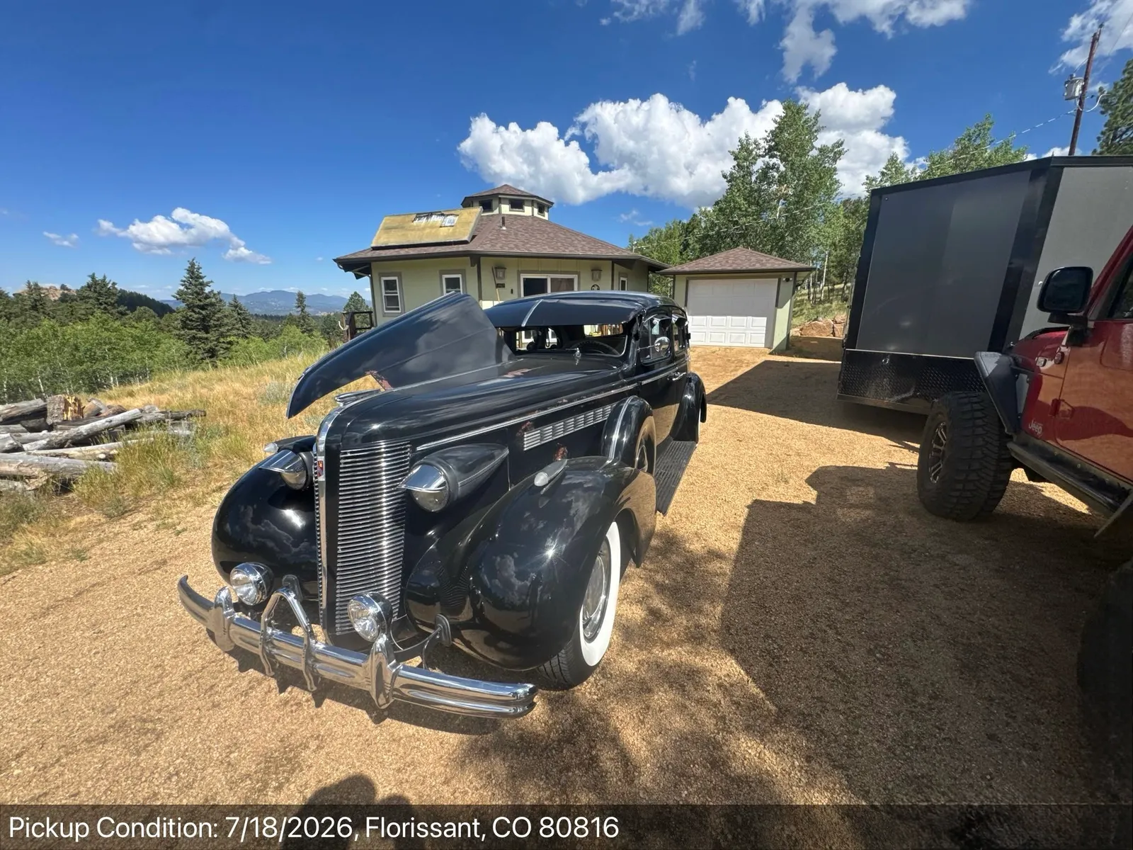 A black 1930s Buick sedan staged beside a small enclosed trailer towed by a Jeep, at a mountain property in Florissant, Colorado