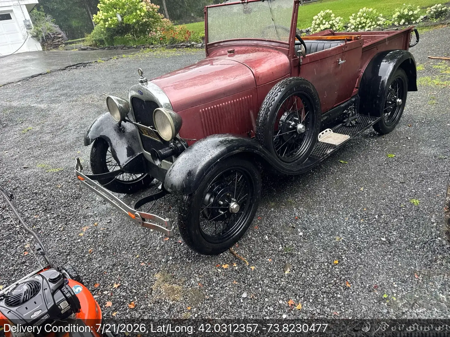 A maroon late-1920s Ford Model A roadster pickup delivered in the rain on a gravel driveway, wire wheels and original body wet from the storm